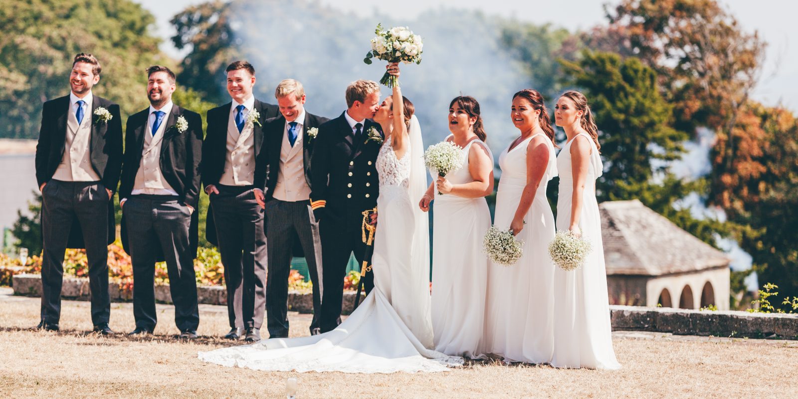 Bride and groom kissing on top lawn with bridesmaids and groomsmen in a line